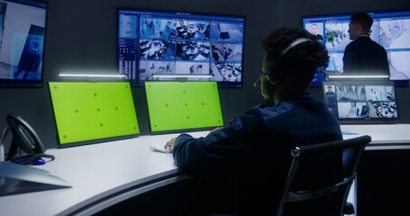 Mixed race officers, operators control security cameras. Employees work in monitoring center. Big digital screens showing surveillance CCTV cameras video footage. Computer monitors with green screen.