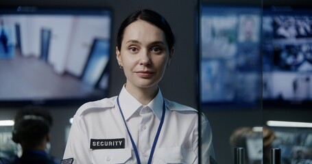 Female security worker in police surveillance center looks at camera. Male employees work on background. CCTV cameras footage displayed on big digital screen. Tracking and monitoring system. Portrait.