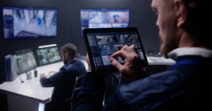 Close up shot of security officer zooming CCTV cameras with AI facial scanning using tablet in police surveillance center. Colleagues work at background. Monitoring system. Concept of social safety.