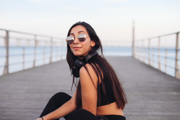 Fototapeta premium Young woman in sunglasses and black headphones enjoying music, sitting by the river side, on a bridge sunset light 