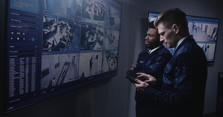Security officer monitors CCTV cameras using tablet and talks with African American colleague in...