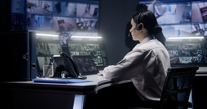 Female security officer in headset zooms and controls CCTV cameras using tablet. Colleagues work at background. Multiple big screens on the wall showing CCTV footage. Monitoring. Social safety concept