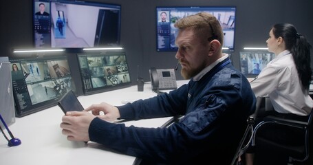 Male security operator in headset controls CCTV cameras with AI facial recognition in police monitoring center. Tablet, PC monitors and big digital screens showing security cameras footage. Tracking.