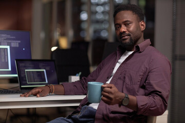 Portrait of African American programmer looking at camera while sitting at his workplace with coffee