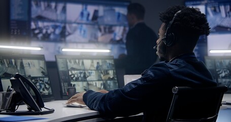 African American male operator in headset monitors security cameras with AI face scanning in police surveillance room. Multiple big screens with CCTV footage at background. Concept of social safety.