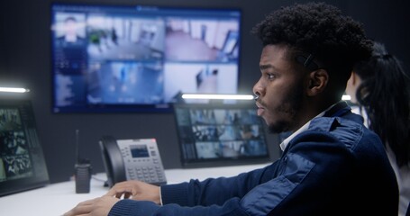 African American officer controls CCTV cameras with AI face scanning in police monitoring room. Female operator works at background. Big screens on the wall showing security cameras footage. Close up.