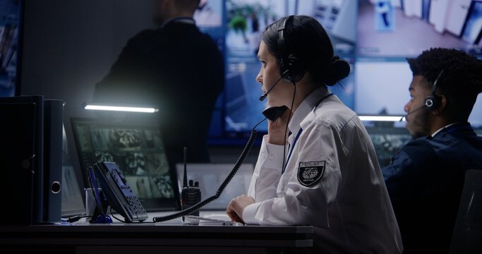 Female security officer works on computer in police monitoring center. Multi ethnic workers control surveillance CCTV cameras with tracking AI face recognition system on tablet and big digital screen.