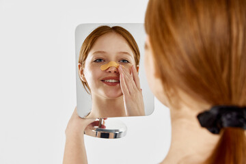 Happy, young, beautiful girl with bandage on her nose after rhinoplasty looking at mirror against...