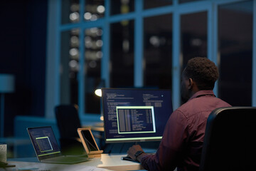 Rear view of African American programmer working with computer codes on computer till late night in...
