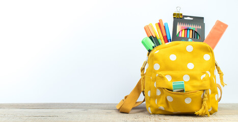 Yellow backpack with white polka dots with different colorful stationery on table. White...
