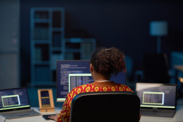 Rear view of African American woman working on computers at her workplace in IT office till late night