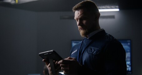 Male security officer controls CCTV cameras in police surveillance center, uses tablet. Multiple...
