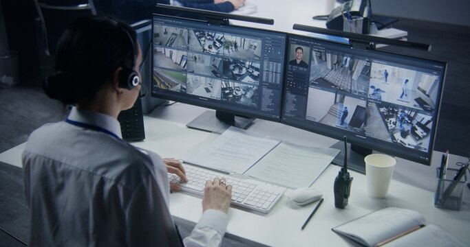 Security officer in headset controls CCTV cameras with AI facial recognition on PC. Female employee works in security control center. Computer monitors showing surveillance cameras video footage.