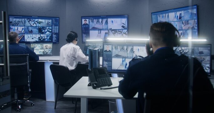 Diverse team of officers watch CCTV cameras with AI facial recognition. Employees work in security control center. Computer monitors and big digital screens showing surveillance cameras video footage.