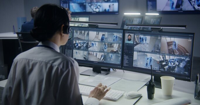 Security officer in headset controls CCTV cameras with AI facial recognition on PC. Female employee works in security control center. Computer monitors showing surveillance cameras video footage.