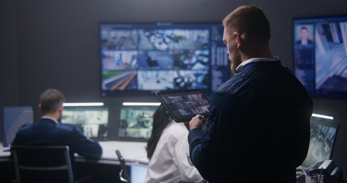 Male officer controls security cameras in police surveillance center, uses tablet. PC monitors and multiple big screens on the wall with CCTV cameras footage. Security operators working at background.