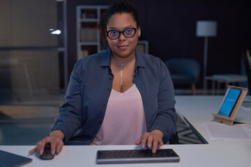Portrait of African American woman in eyeglasses looking at camera while working on computer till...
