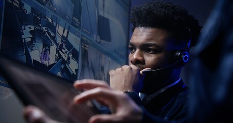 Close up shot of African American male operator in headset talk with colleague and monitor CCTV...