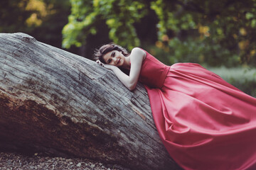 Beautiful young woman in red dress leans on tree trunk