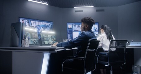 African American male officer types keyboard and uses walkie talkie. Multi ethnic employees in police control center. Big digital screens and PC monitors with surveillance CCTV cameras video footage.