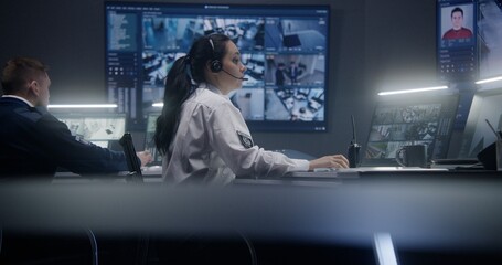 Female operator types keyboard and monitors security cameras with AI facial recognition in police surveillance room. PC monitors and big digital screens on the wall with displayed CCTV cameras view.