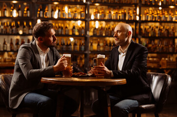 adult friends sitting with beer mugs in a pub. Two cheerful guys drink draft beer, celebrate meeting and talk