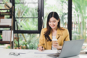 Obraz premium Happy Business woman entrepreneur sit and holding cup of coffee and working with report and laptop at office desk. Portrait of beautiful smiling young businesswoman working at modern work station.