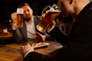 adult friends sitting with beer mugs in a pub. Two cheerful guys drink draft beer, celebrate meeting and talk