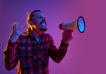 Portrait of angry emotional man shouting in megaphone with annoyance against purple studio...