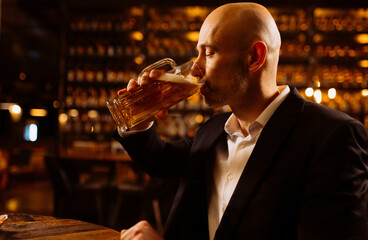 A man drinks beer. Side view of handsome bald man drinking beer in evening pub. Businessman drinks beer after work.