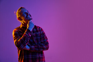 Portrait of happy smiling man with thoughtful face against purple studio background in neon light. Success, ideas. Concept of human emotions, facial expression, lifestyle, business