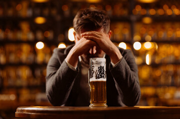 Sad young man in a suit looking at a glass of beer while sitting at a table in a pub. A man drinks after work in a pub.