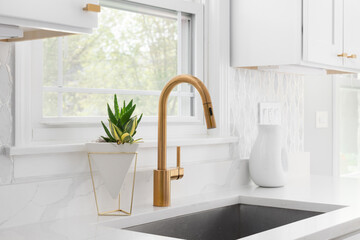 A kitchen sink detail with a gold faucet, marble tile backsplash, and white cabinets.