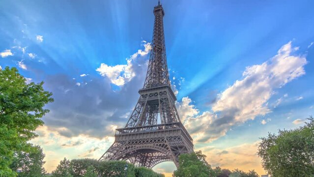 Sun behind the Eiffel tower with warm rays of light in clouds during sunset timelapse hyperlapse. It is one of the most recognizable landmarks in the world. Blue cloudy sky at sunny evening