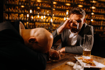 A young man in a suit sleeps near a glass of whiskey and beer on a table in a pub, another man drinks alcohol