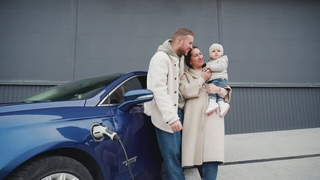 Happy family having fun together near blue electric car. Mom, dad and little daughter together.