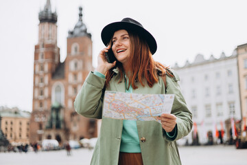 Fototapeta premium Beautiful stylish woman in hat walking on Market Square in Krakow on autumn day and holding mobile phone and map. Phone Communication. Urban lifestyle concept. Check social networks, booking hotel