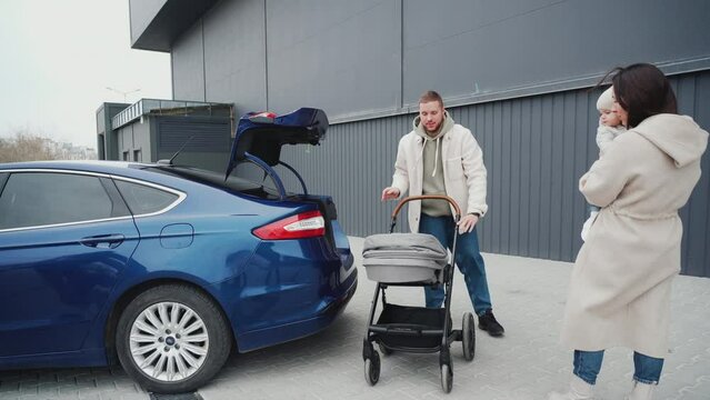 A young father takes a baby carriage out of the trunk of a blue electric car. A father makes a baby carriage for his little daughter.