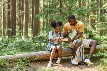 African American dad having snack together with his daughter while they sitting on log after hiking