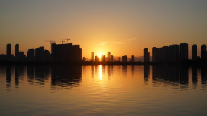 The beautiful sunset view with the buildings' silhouette and orange color sky mirrored in the peaceful lake in the city