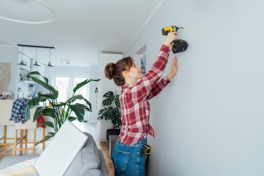 Back view young brunette woman screwing screw into wall with electrical screwdriver. Girl wants to put a picture on the wall. Housekeeping work. Doing repairs herself, DIY, gender equality concept.