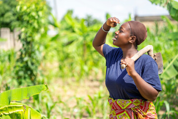image of african lady in a green field holding a farm tool- black lady in a farm looking to the sky with hole on the shoulder
