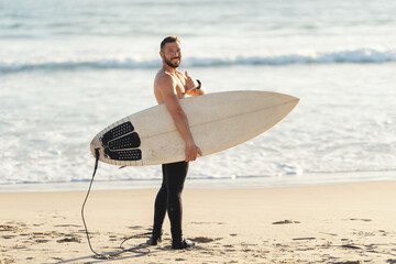 An athletic man surfer with naked torso standing by the sea and showing a thumb up looking in the camera