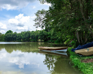 An unused wooden boat stands by the lake at the natural park in spring and summer seasons with cloudy blue sky. Boat floating on the reflected river among green trees in serenity day, calm nature.