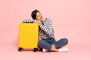 Happy Young Asian traveler woman sitting on floor with yellow suitcase bag isolated on pink background, Tourist in holidays vacation concept