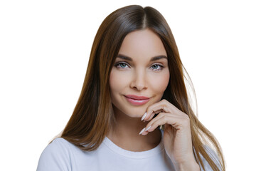 Charming good looking millennial woman keeps hand near face, has healthy glowing skin, dark hair, blue eyes, dressed in casual wear, isolated over brown background. Human face expressions concept