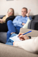 Hand of psychologist holding pen ready to make notes against blurred couple arguing on sofa. Man and woman family therapy session