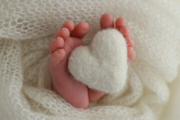 Knitted white heart in the legs of a baby. Macro photography. The tiny foot of a newborn baby. Soft feet of a new born in a white wool blanket. Close up of toes, heels and feet of a newborn.