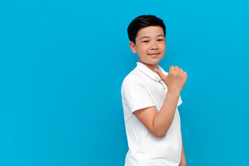 little asian boy in white t-shirt points back on blue background, korean child advertises copy space