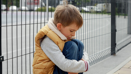 Portrait of little boy crying while sitting alone next to metal fence on street. Child depression,...